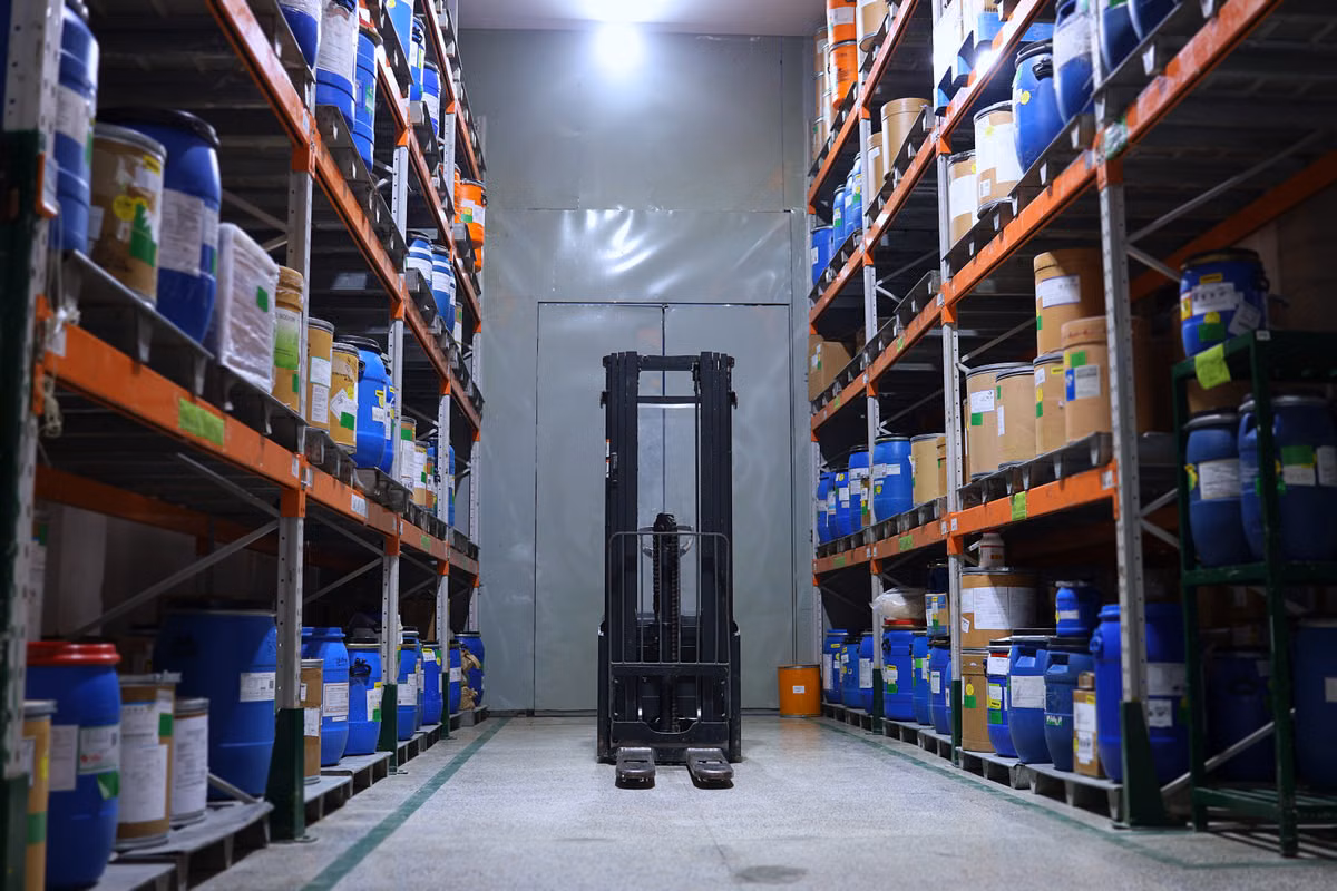 An unoccupied reach truck parked in an aisle between warehouse racking stacked with drums and containers.