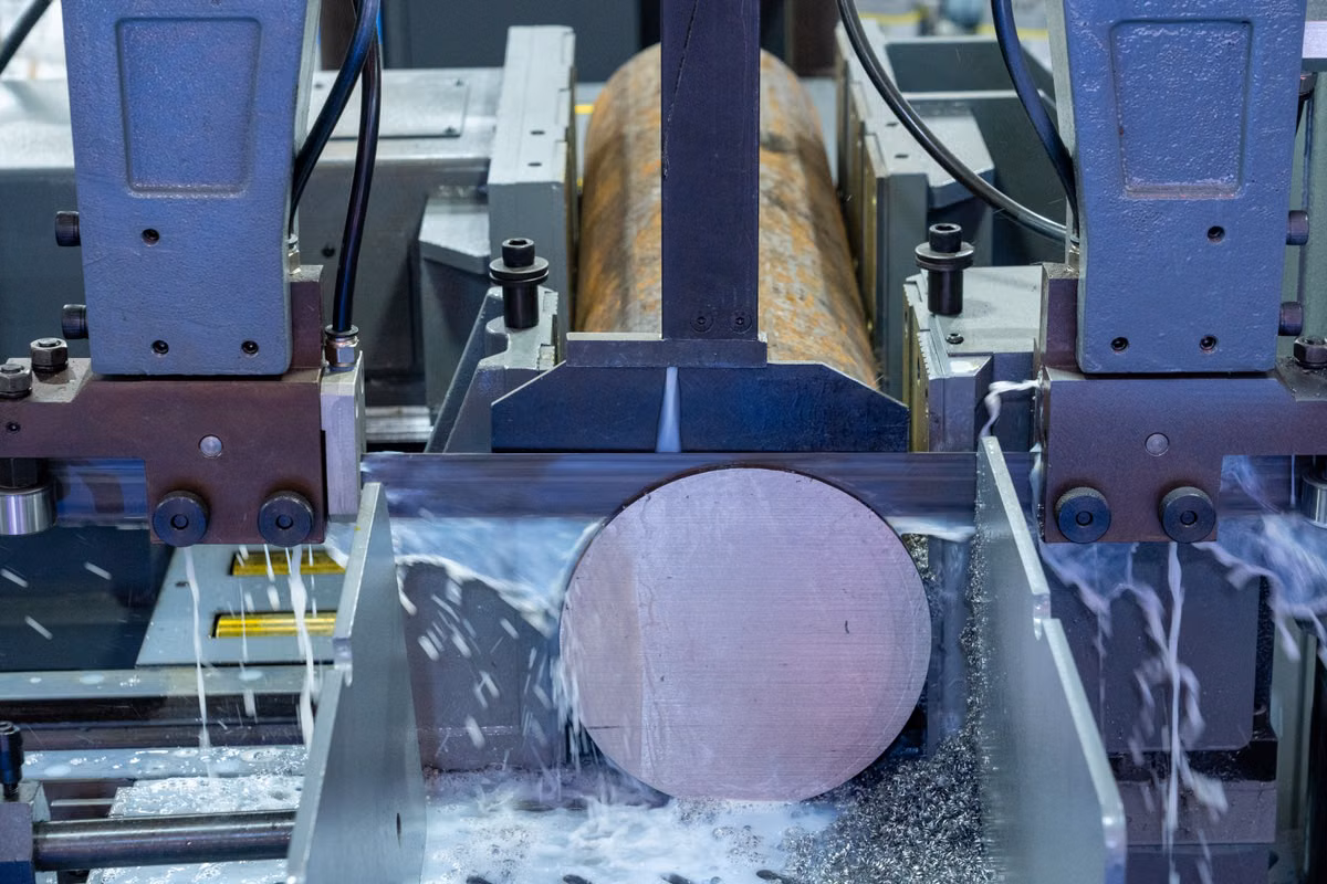Close-up of an automatic band saw cutting through a round metal bar, with coolant spraying around the blade and clamps.