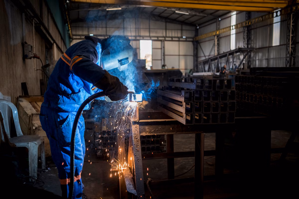 A welder in blue overalls, gloves and a welding helmet welds steel box sections inside a fabrication workshop as sparks fly.